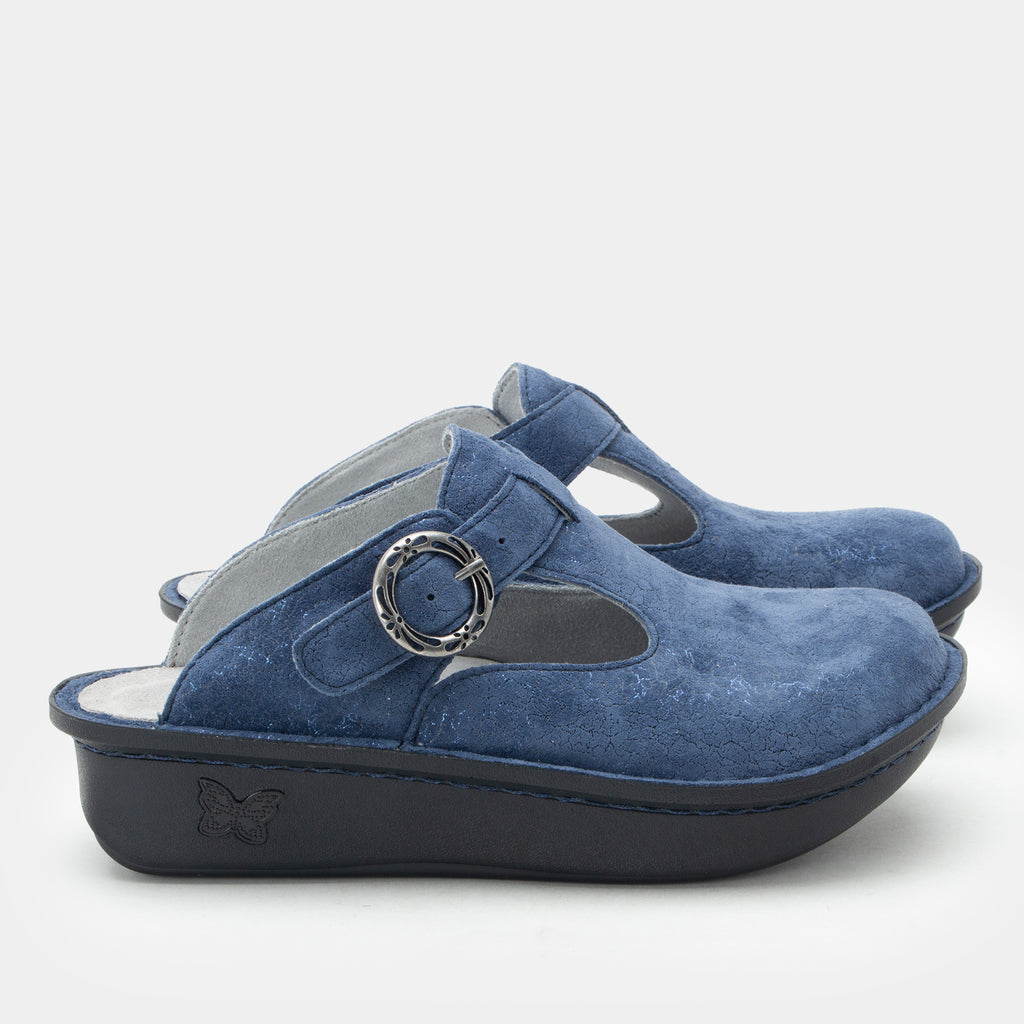 Shimmer Blue  suede slip-on clogs shown with a dark sole, featuring a circular metal buckle detail on the strap and a small butterfly logo on the heel. ALG-6559