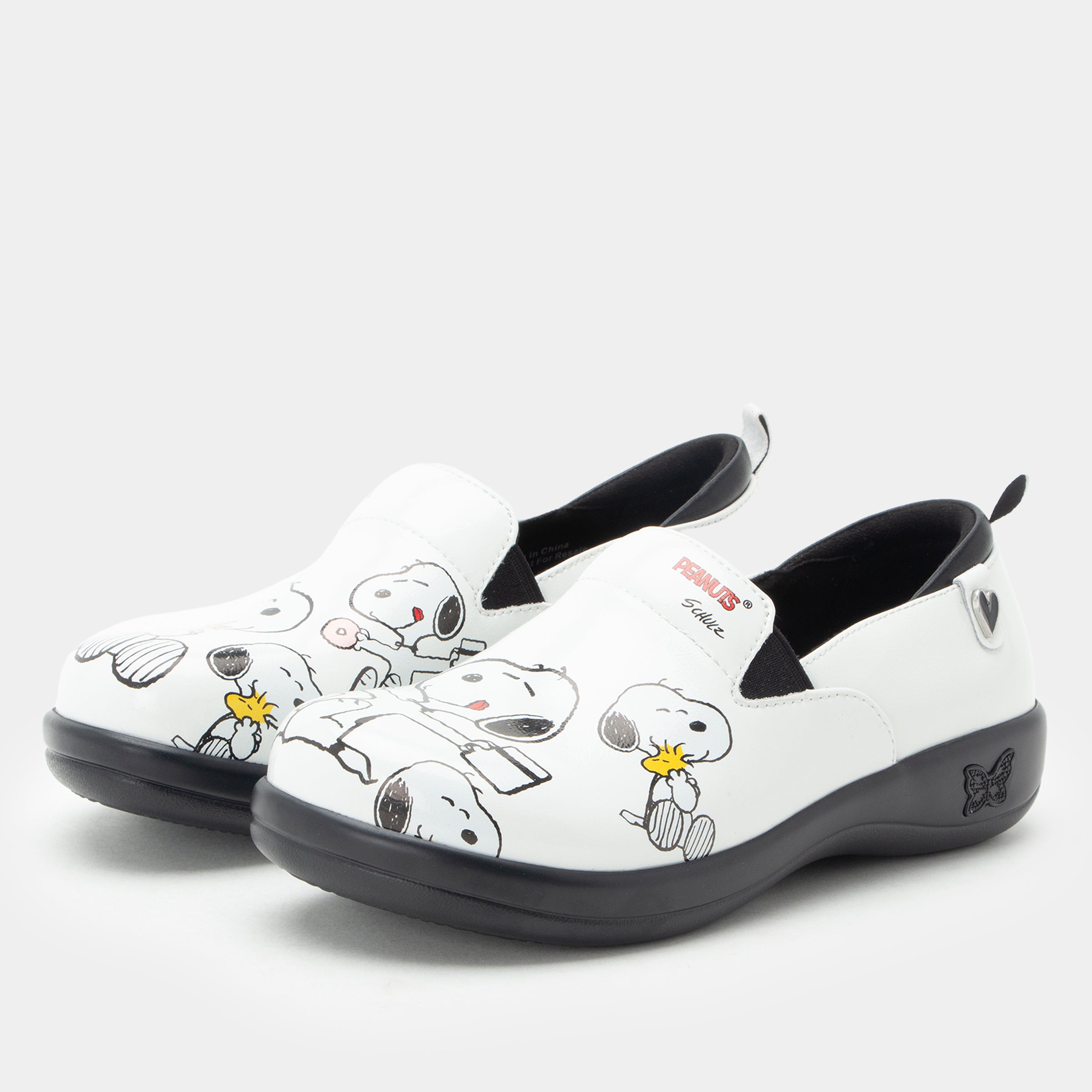 A pair of Alegria x Peanuts® white slip-on shoes featuring a black sole and cartoon Peanuts® characters print on the leather uppers. KEL-SNP19