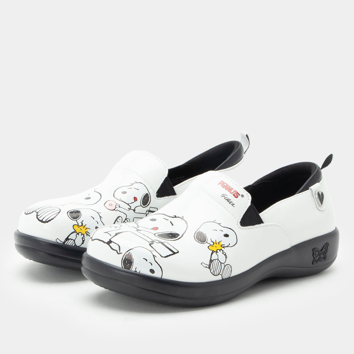 A pair of Alegria x Peanuts® white slip-on shoes featuring a black sole and cartoon Peanuts® characters print on the leather uppers. KEL-SNP19