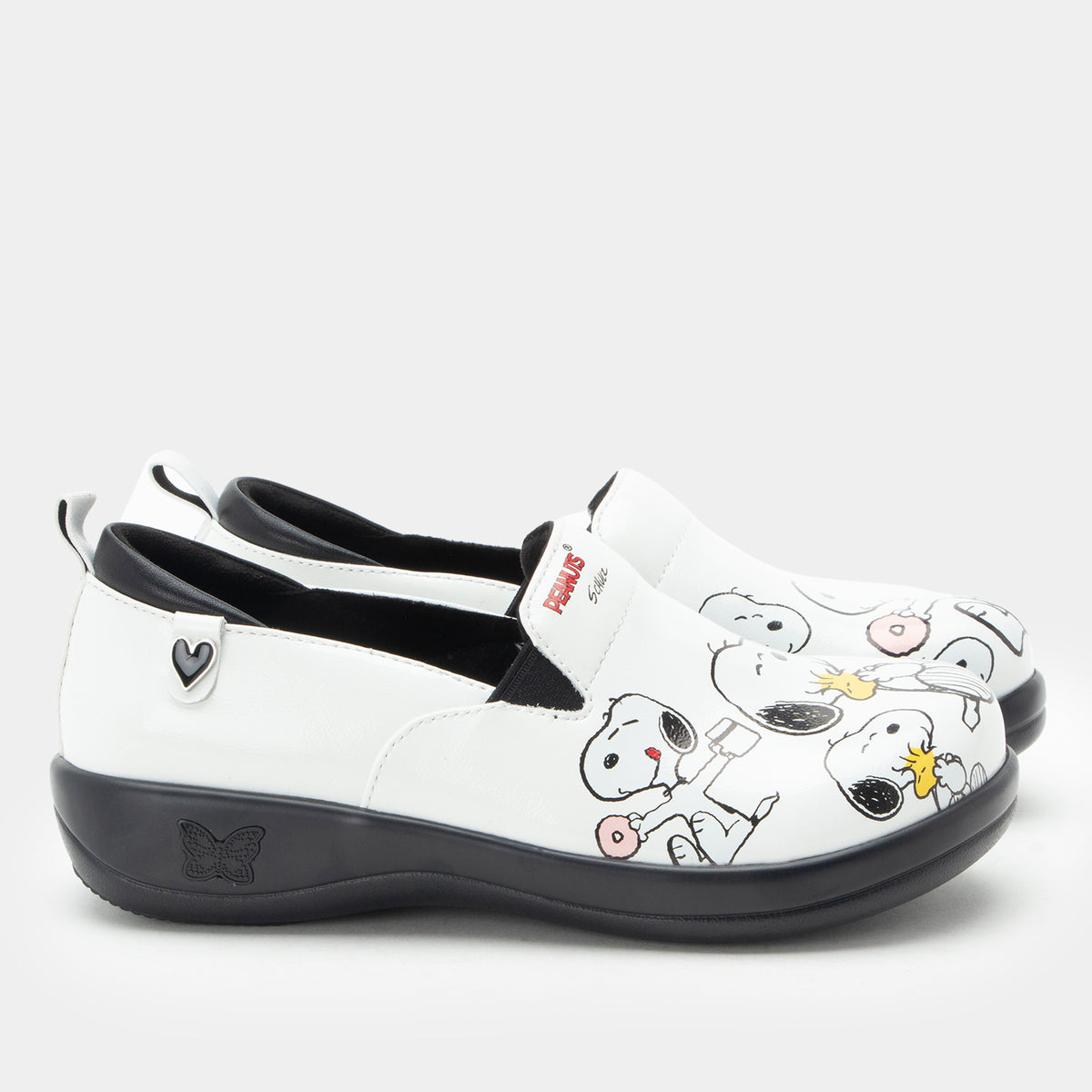 A pair of Alegria x Peanuts® white slip-on shoes featuring a black sole and cartoon Peanuts® characters print on the leather uppers. KEL-SNP19
