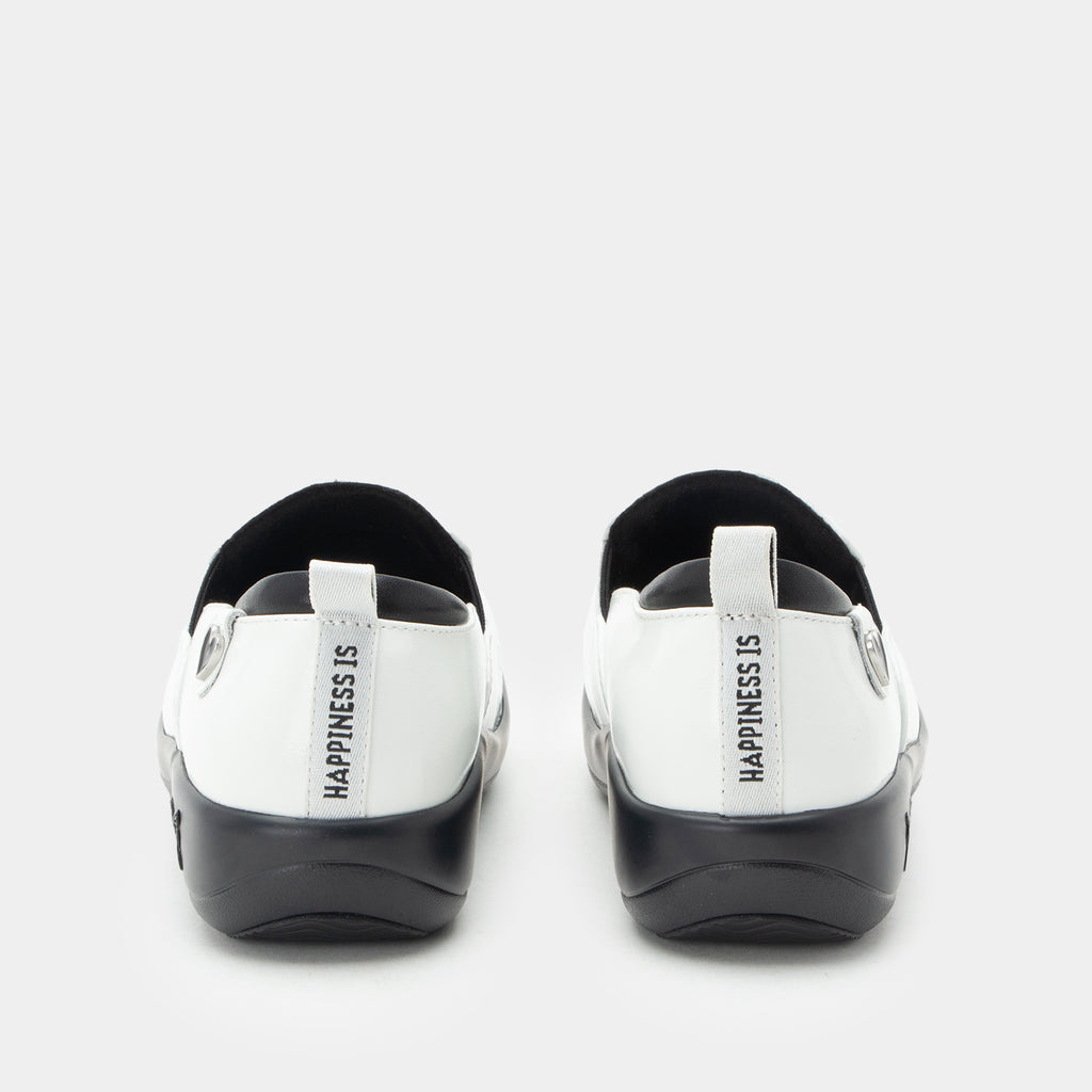 A pair of Alegria x Peanuts® white slip-on shoes featuring a black sole and cartoon Peanuts® characters print on the leather uppers shown from the back. The heel area features a white pull tab with the text 'HAPPINESS IS' printed vertically in black. KEL-SNP19