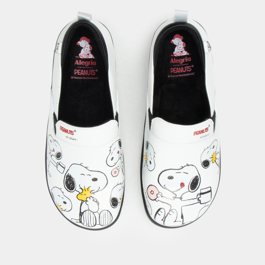 A pair of Alegria x Peanuts® white slip-on shoes featuring a black sole and cartoon Peanuts® characters print on the leather uppers shown from the top view with black interiors and red 'Alegria PEANUTS' branding. KEL-SNP19