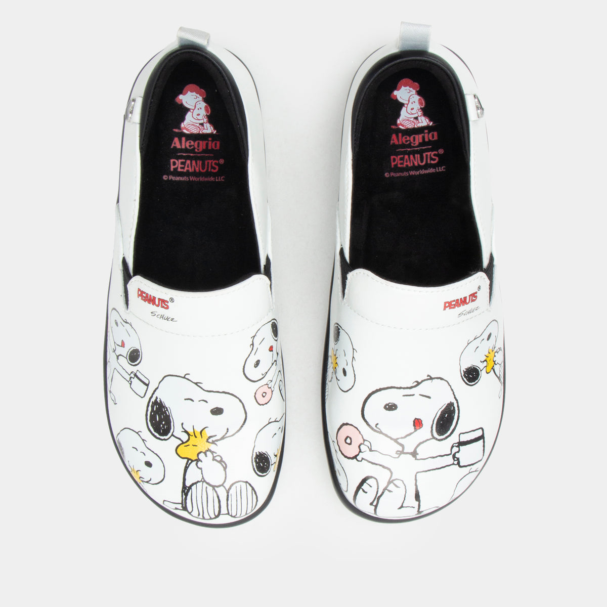 A pair of Alegria x Peanuts® white slip-on shoes featuring a black sole and cartoon Peanuts® characters print on the leather uppers shown from the top view with black interiors and red 'Alegria PEANUTS' branding. KEL-SNP19