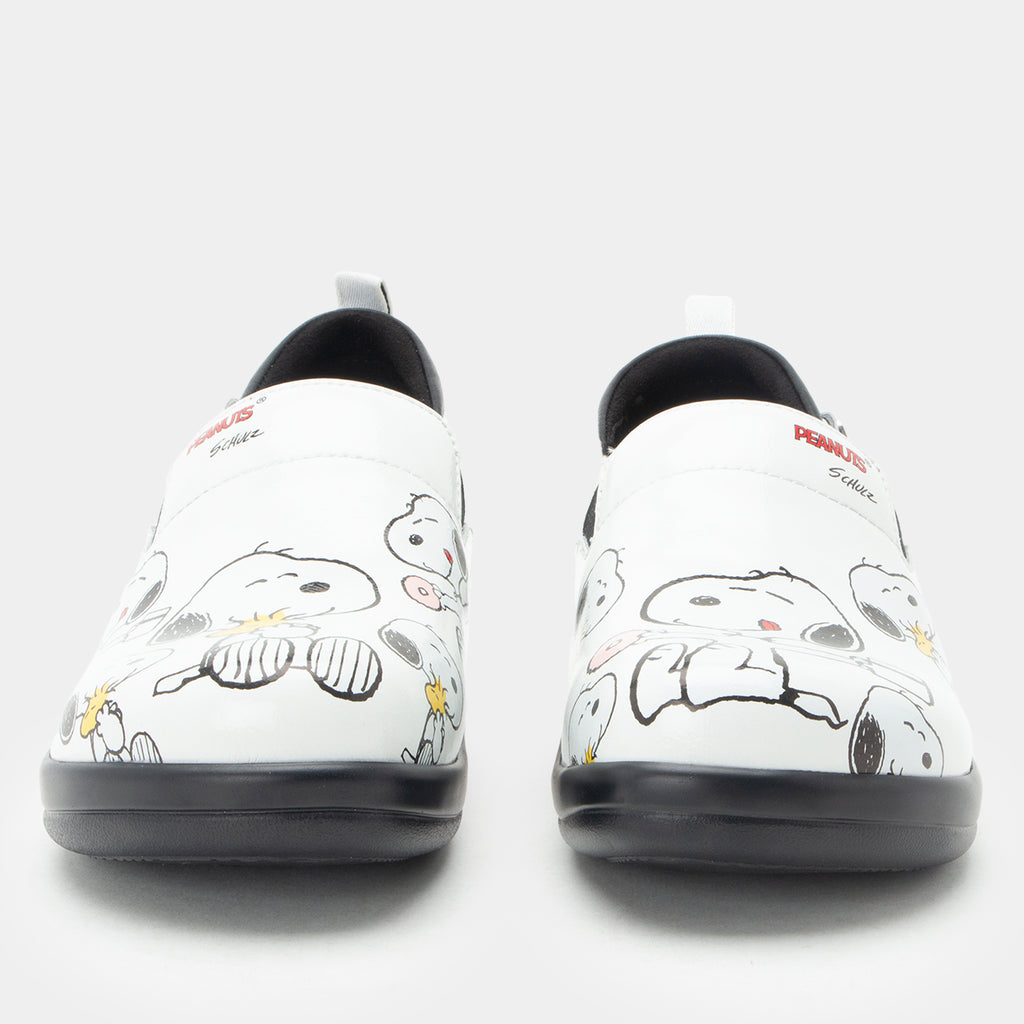 A pair of Alegria x Peanuts® white slip-on shoes featuring a black sole and cartoon Peanuts® characters print on the leather uppers shown from a front-facing angle. KEL-SNP19