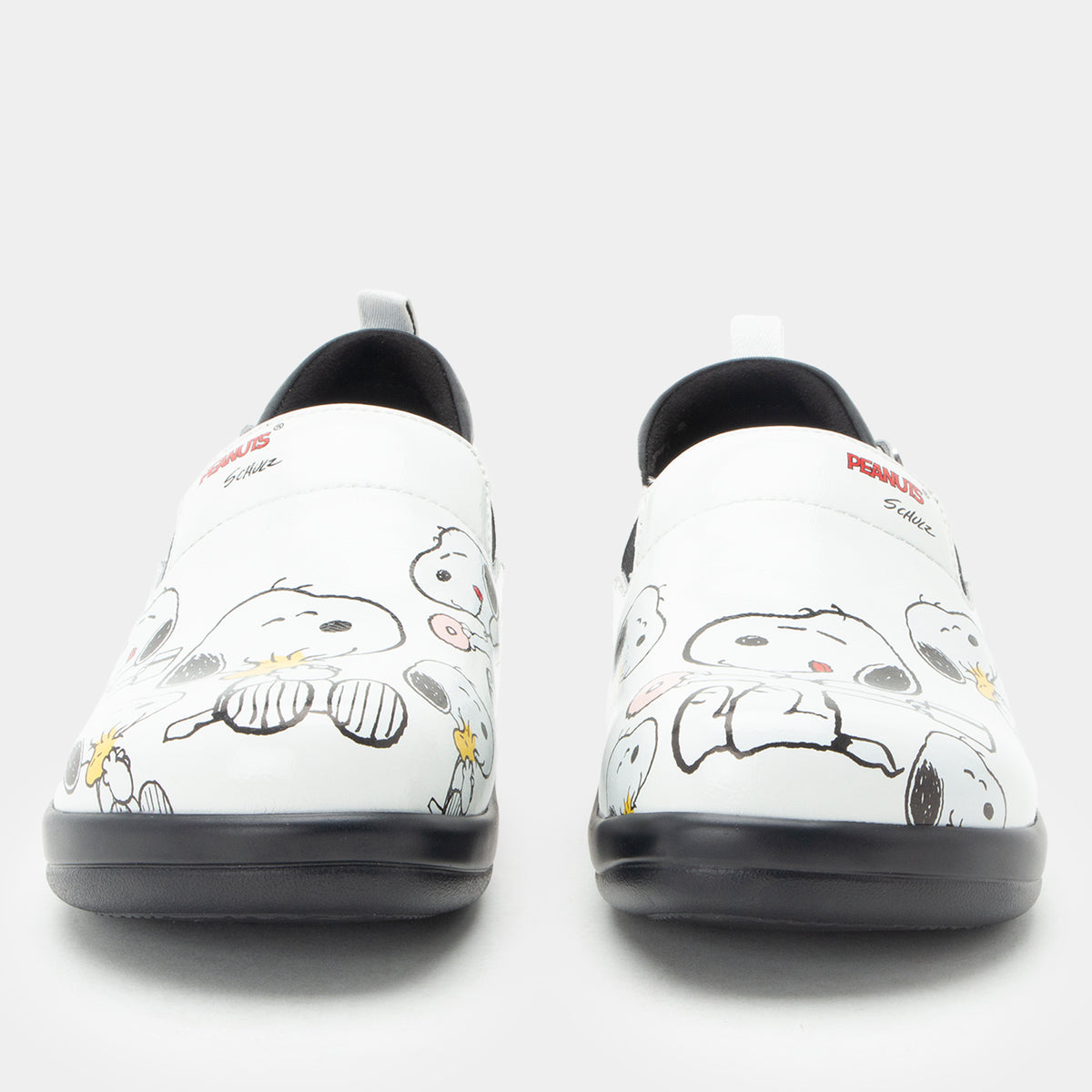 A pair of Alegria x Peanuts® white slip-on shoes featuring a black sole and cartoon Peanuts® characters print on the leather uppers shown from a front-facing angle. KEL-SNP19